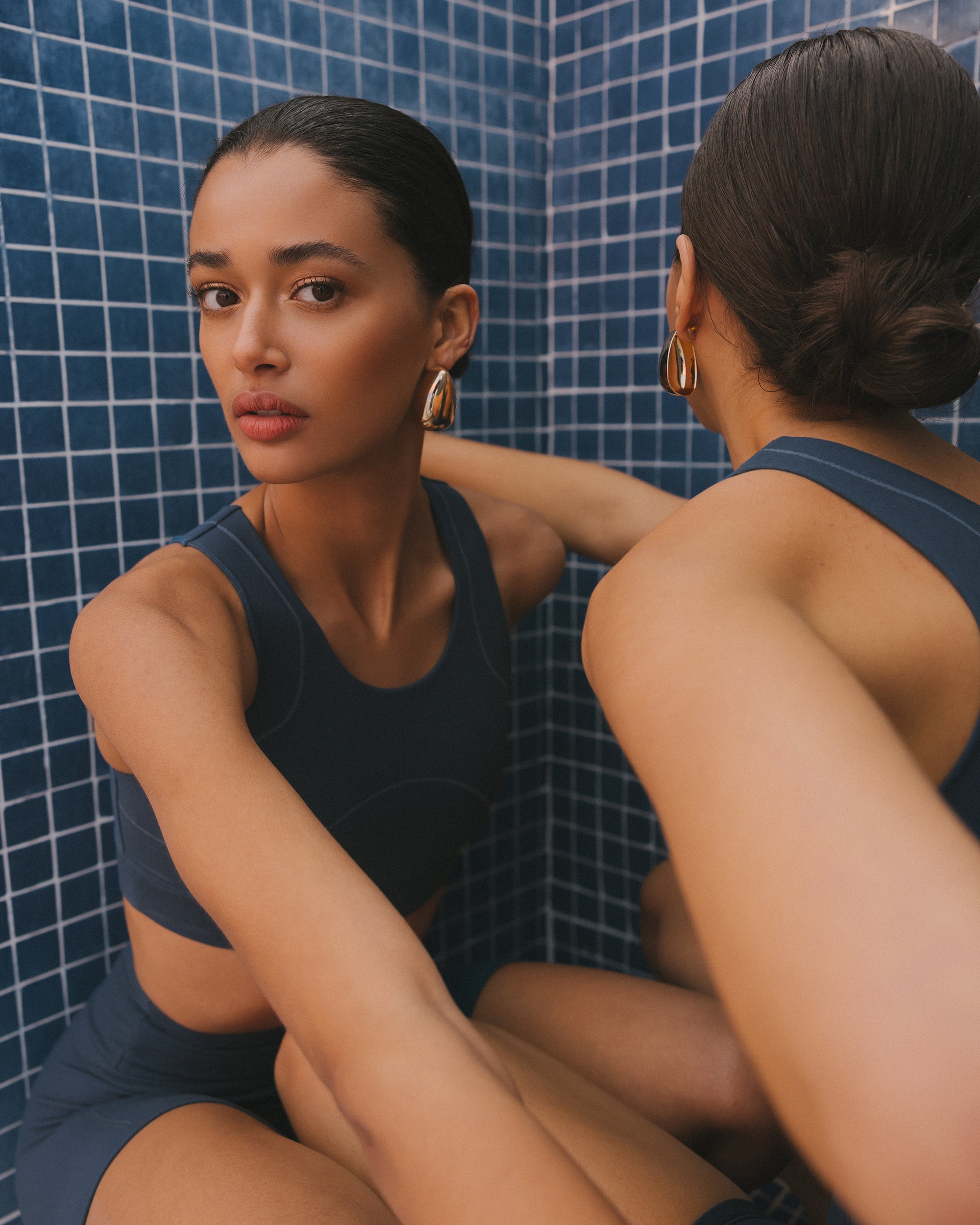 A woman in dark blue activewear gazes intently, with another turned away, in a room with blue mosaic tiles, highlighting a moment of focused tranquility.