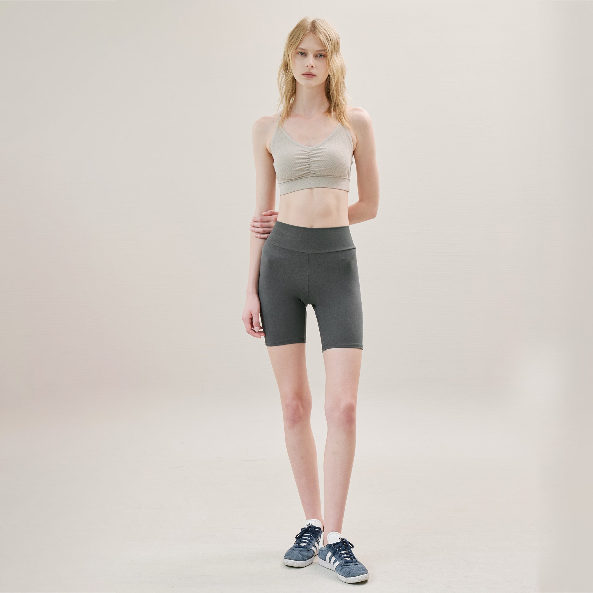 Woman wearing a beige sports bra and gray shorts on a plain background
