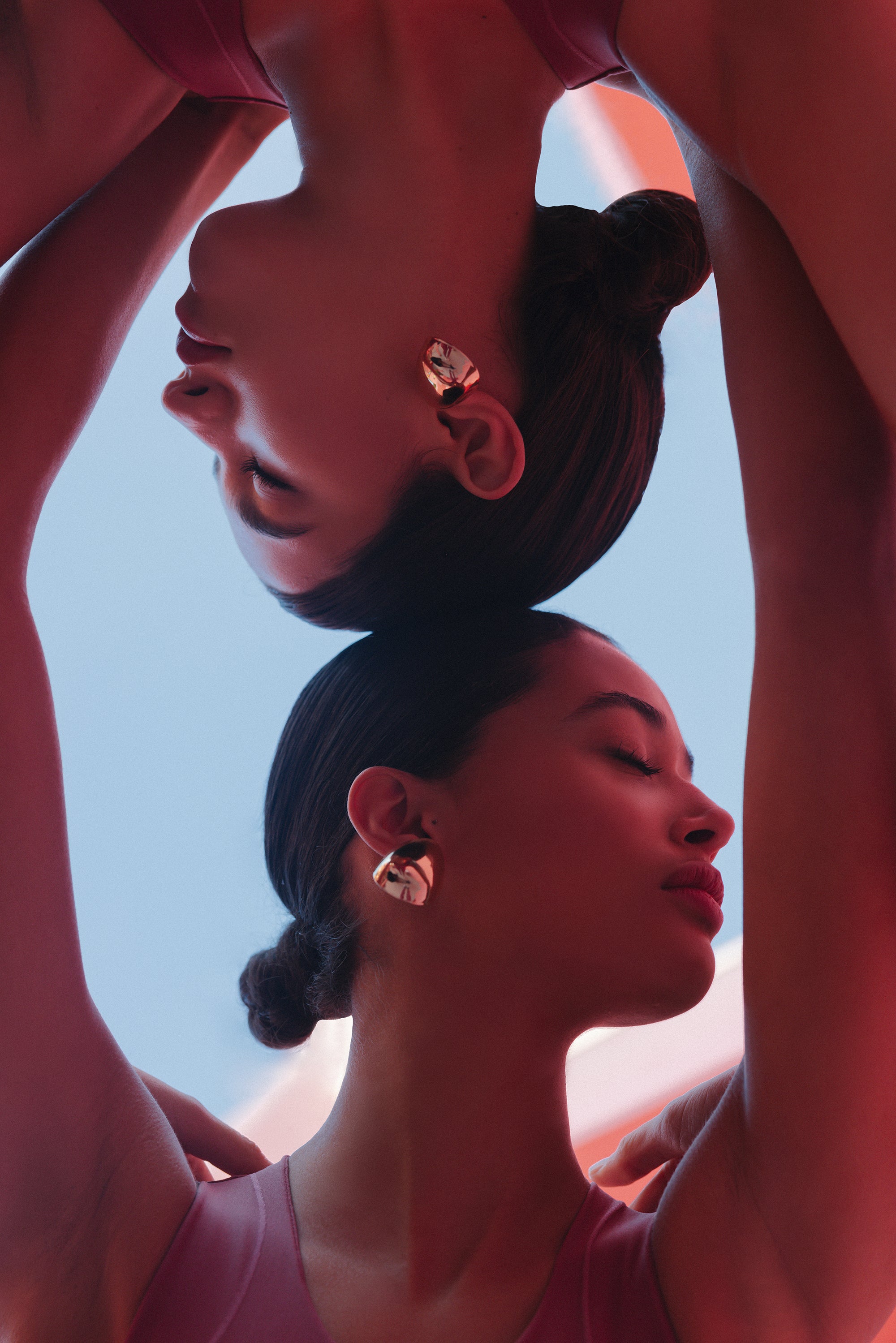 Two women, adorned in mauve activewear, form an archway with their arms and heads touching in a mirrored, symmetrical fashion, evoking a sense of connection and continuity.