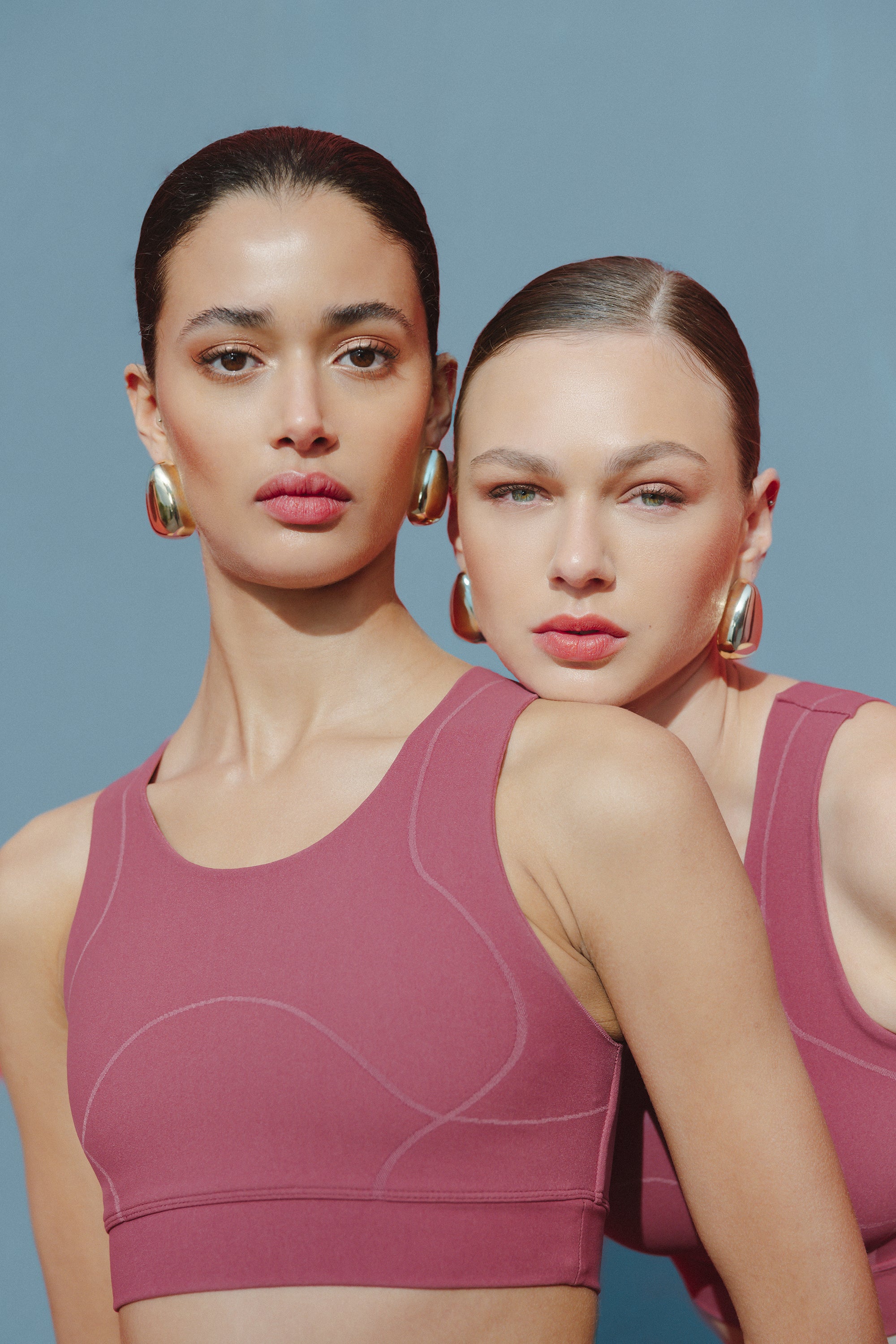 Two models in coordinated mauve activewear tops, with one facing forward and the other in profile, against a calm blue backdrop.