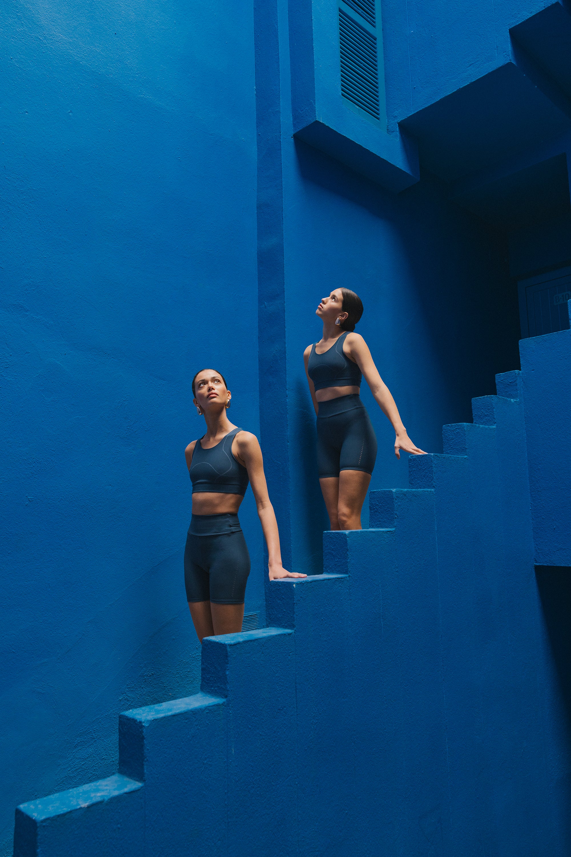 Two women in dark activewear ascend a vibrant blue stairwell, pausing to reflect, embodying strength and serenity.