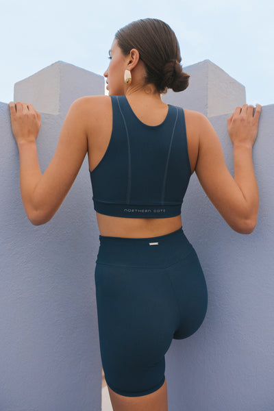 A woman in dark blue activewear leaning against a wall, looking over her shoulder, set against a muted blue background.