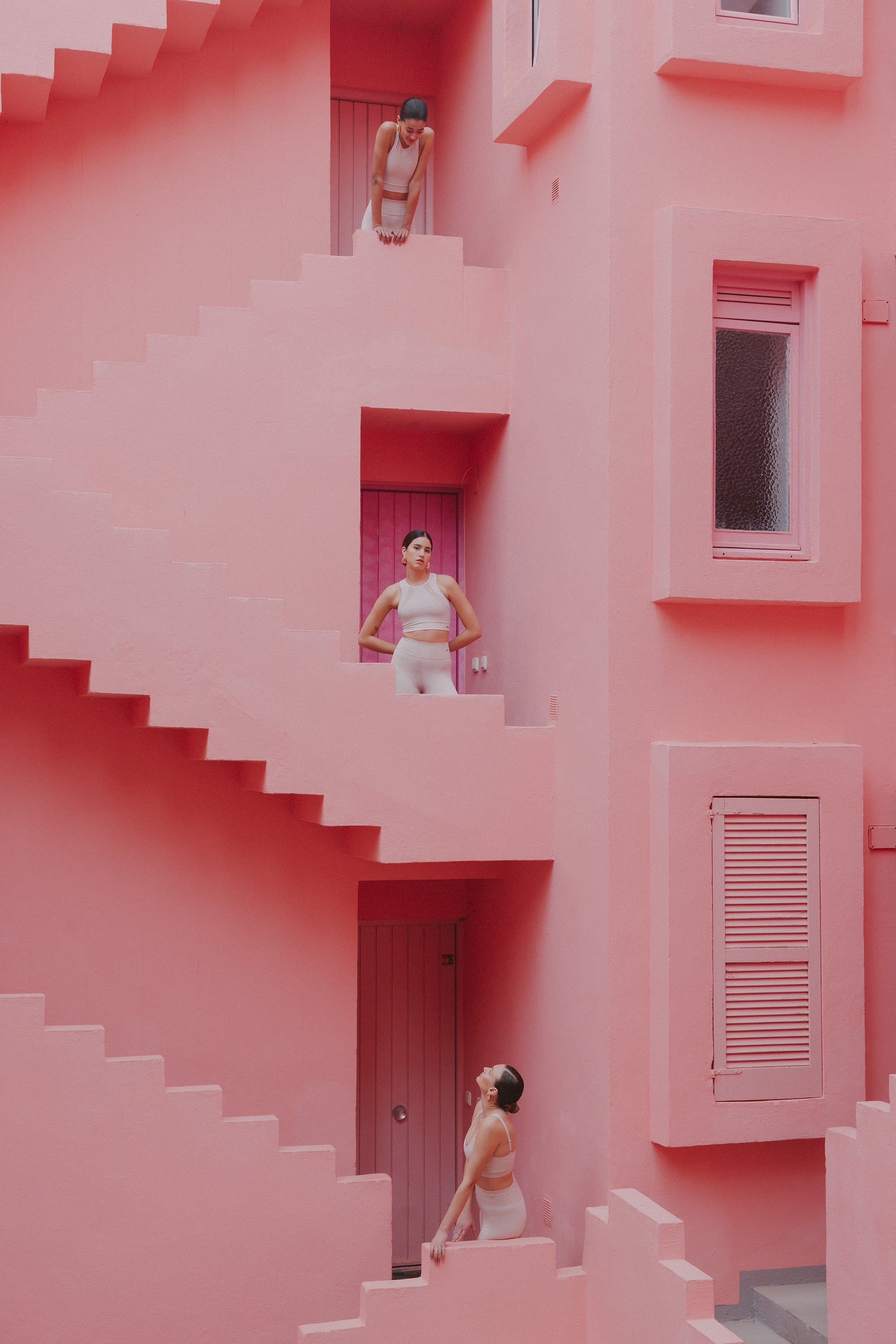 Three women in white activewear positioned at different levels in a stairwell, within a striking pink architectural complex.