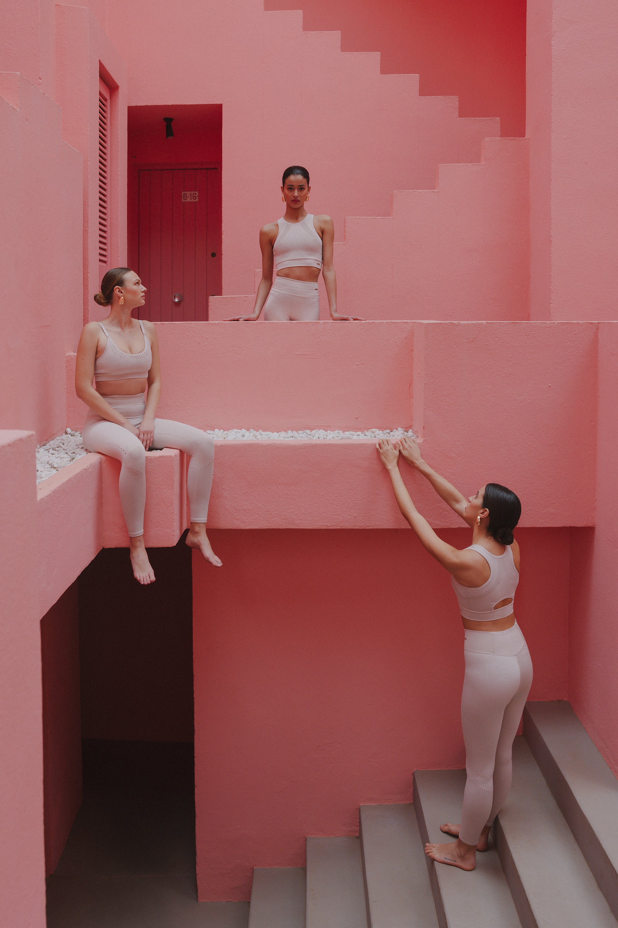 Three women in white activewear engaging in various poses within a pink stairwell, creating a playful and dynamic scene.