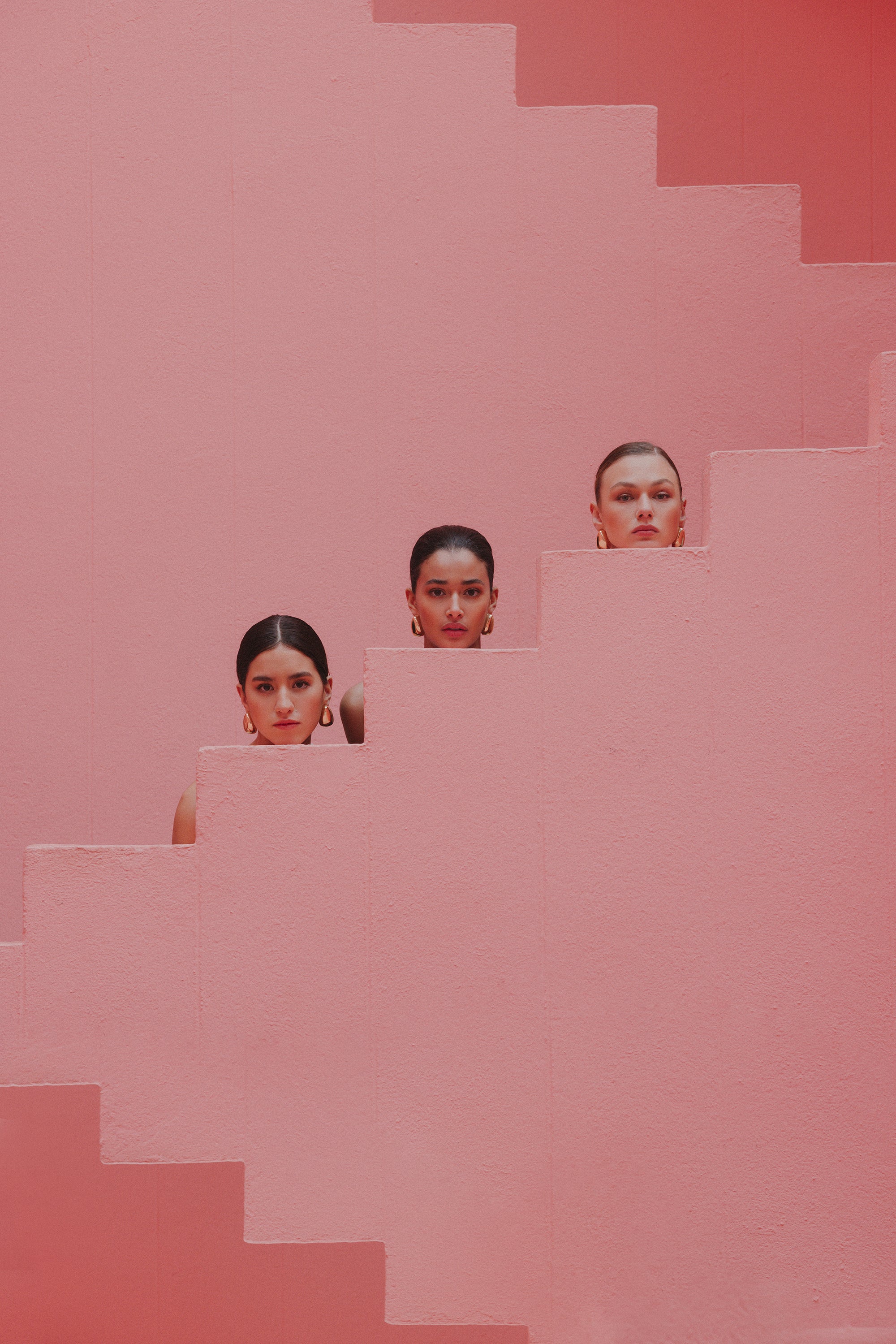 Three women in white activewear peek over the edges of a pink staircase, their faces framed by the geometric shapes of the architecture.