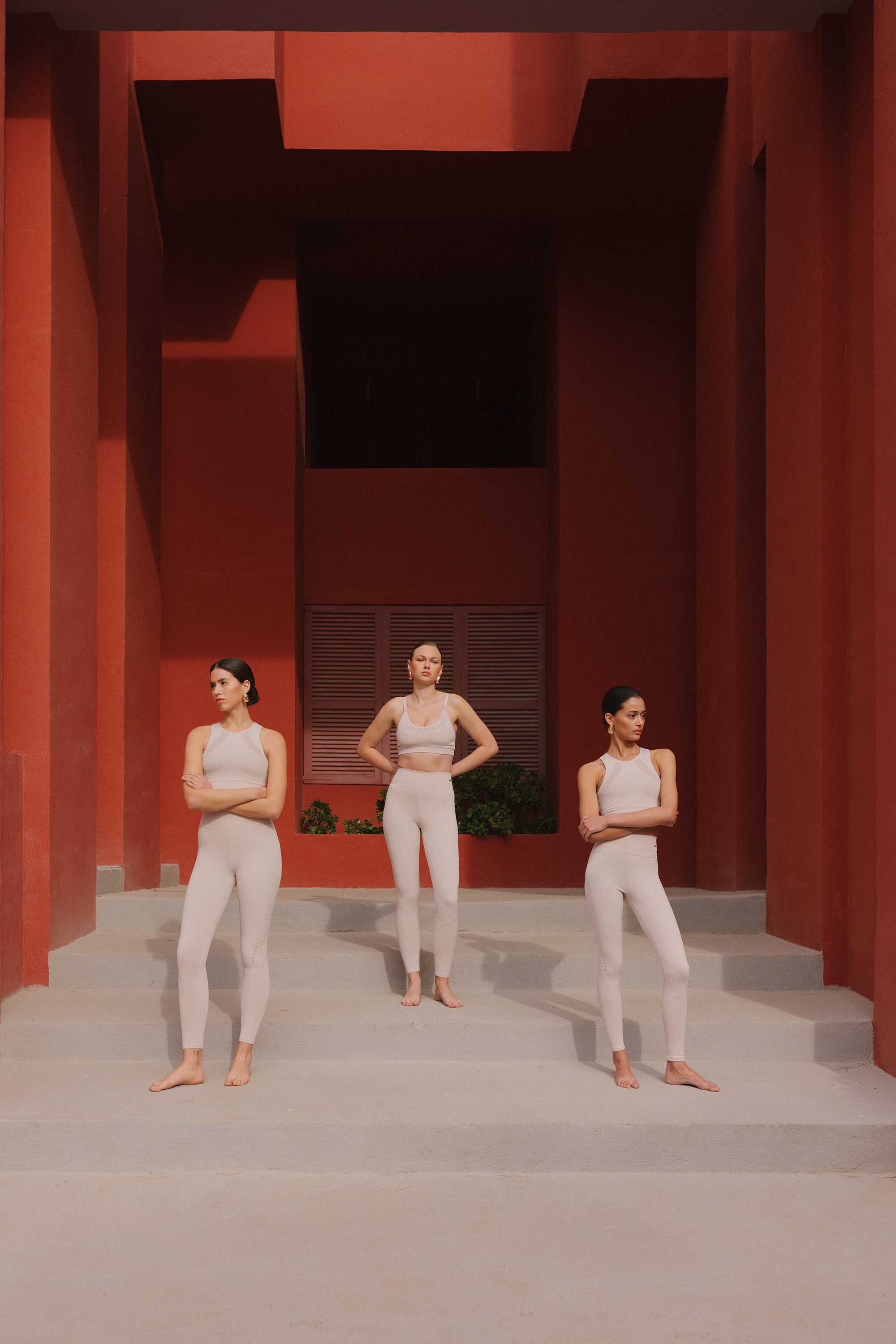 Women in white activewear stand poised on a terracotta stairway, their calm expressions contrasting with the bold architecture around them.