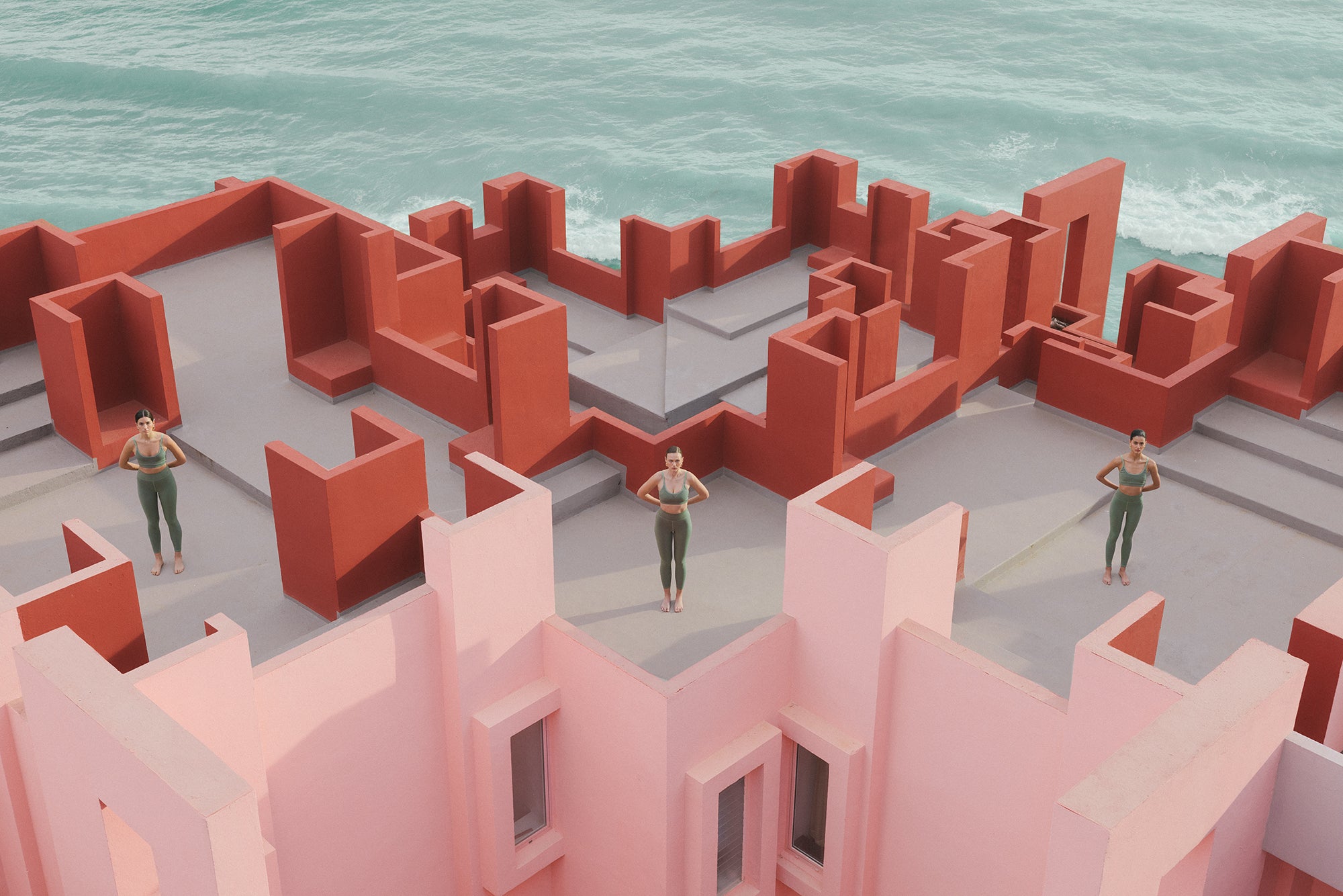 Women in olive activewear stand amid a labyrinth of red walls against the backdrop of a turquoise ocean.