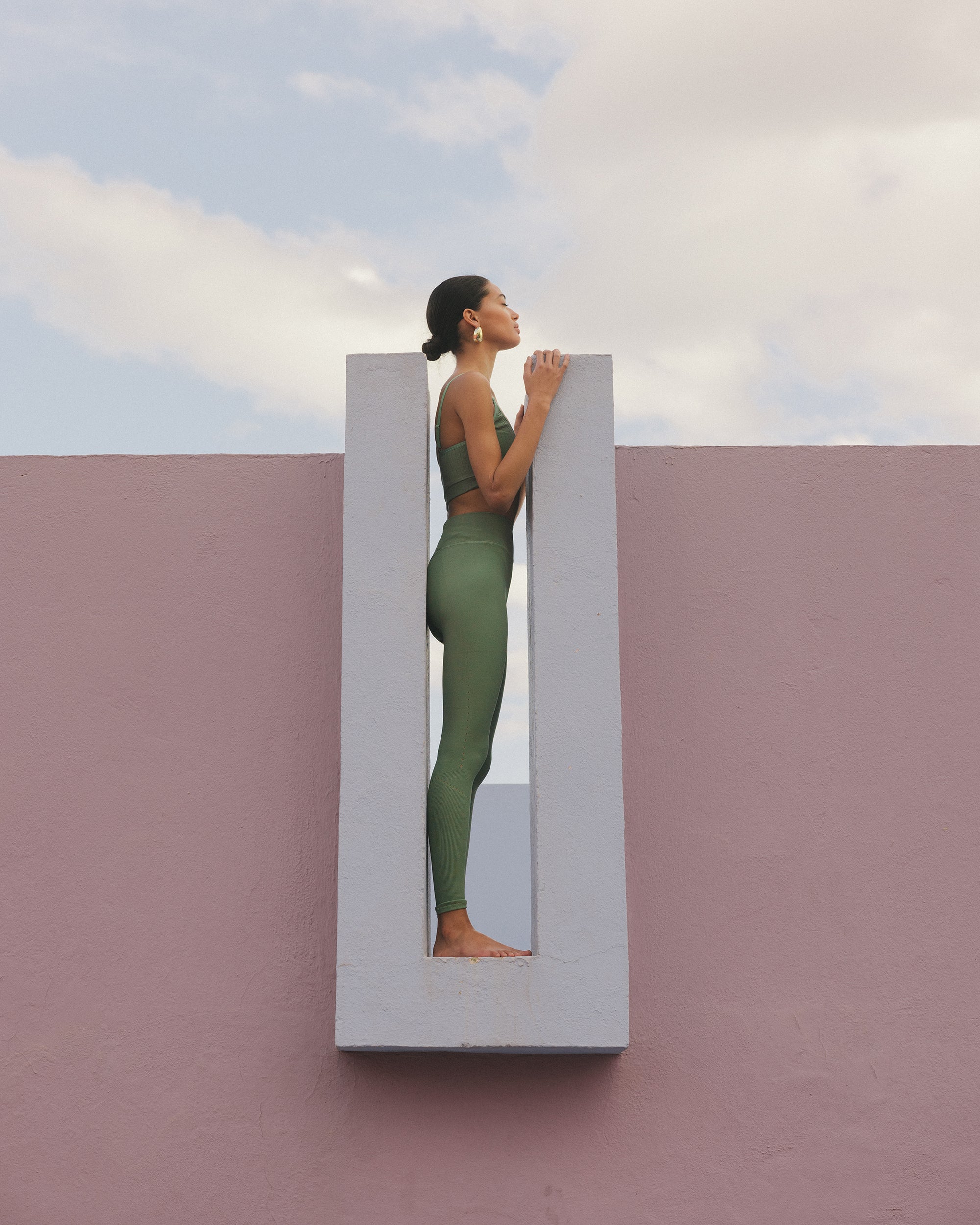 A contemplative woman in olive green activewear stands within a narrow vertical window, with a pastel sky in the background.