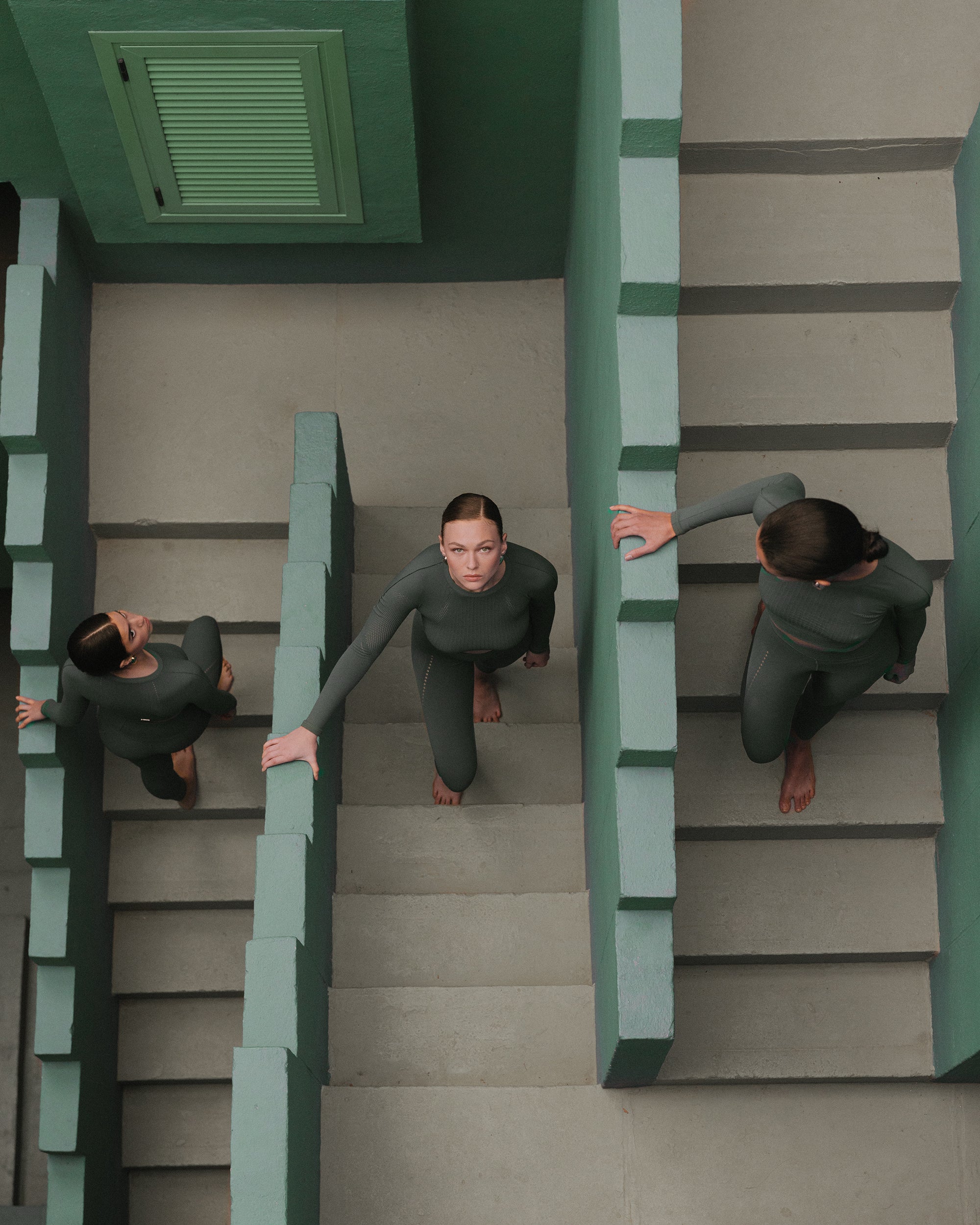 Women in dark green activewear descend a stairway, their figures echoed against the stark geometry of the architecture.