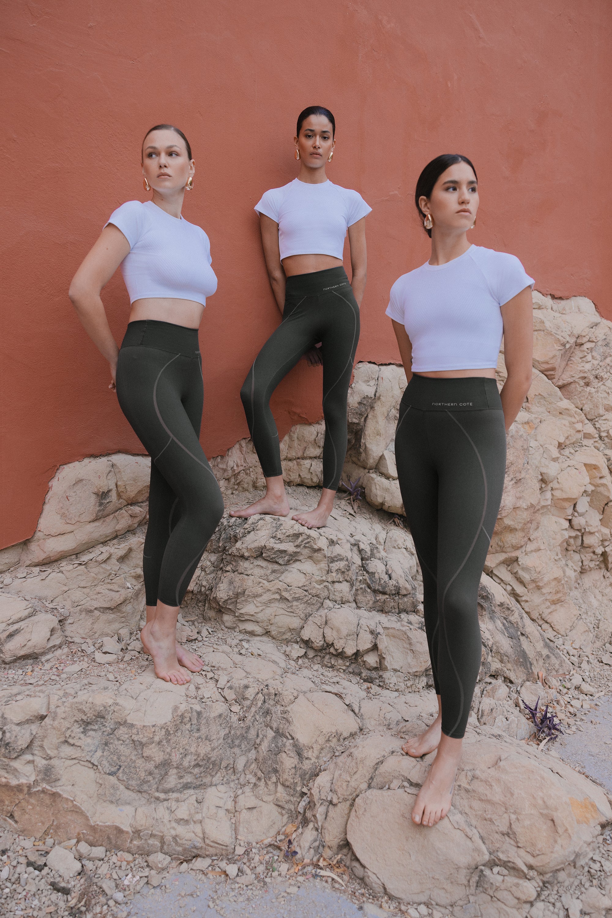 Three women in coordinated dark green leggings and white cropped tops posed on rocky terrain against a coral-colored wall.