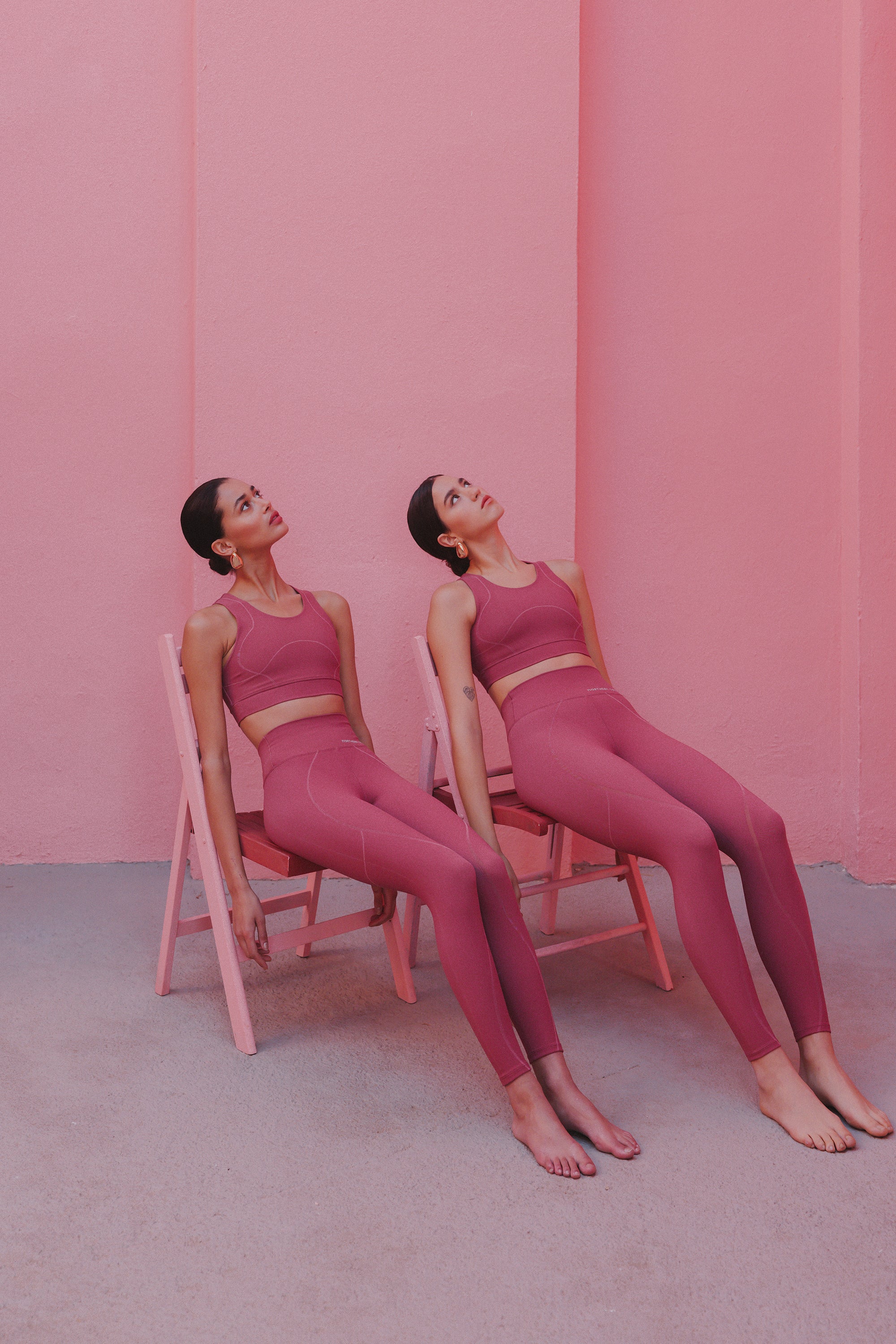 Two women dressed in matching burgundy activewear, lounging on pale pink chairs against a wall of the same color, their poses relaxed and their gazes directed upward.